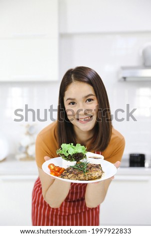 Young female cooking in the kitchen, Asian woman showing steak plate in kitchen. Selective focus. Royalty-Free Stock Photo #1997922833