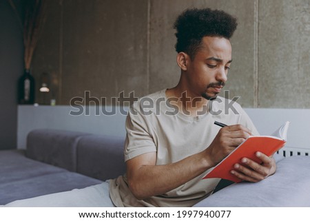 Pensive young poet student african american man 20s wearing beige t-shirt sit on grey sofa indoors apartment writing down poem in notebook diary, resting on weekends staying at home, enjoying hobby. Royalty-Free Stock Photo #1997940077