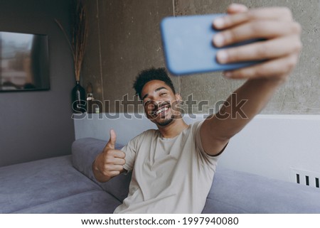 Fun happy young african american man in beige t-shirt sit on grey sofa indoor apartment doing selfie shot on mobile phone post photo on social network show thumb up gesture internet rest stay at home.