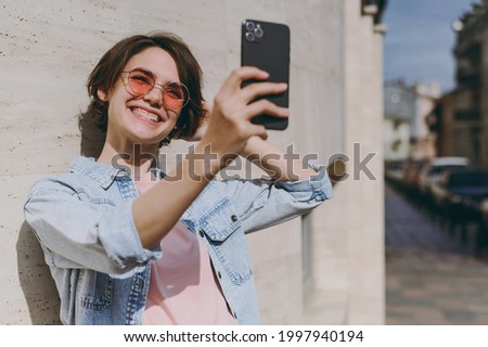 Young smiling caucasian happy fun woman 20s wear jeans clothes glasses do selfie shot on mobile phone post photo on social network outdoors near city street building wall. People lifestyle concept.