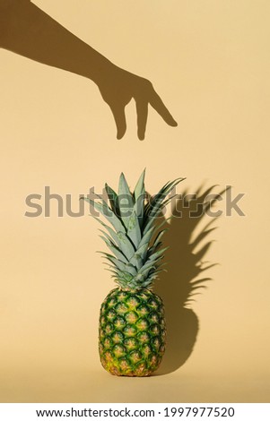 Creative summer vibes concept with fresh raw pineapple and woman’s hand shadow. Aesthetic tropical fruit idea. Minimalistic vertical arrangement. Pastel yellow background.