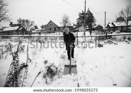 An old woman cleans the snow near home in the village. Black and white photo.