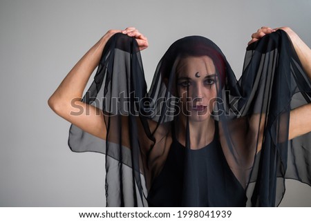 Redhead woman with a black veil on her head in a dark studio. A girl with an earring in her nose smiles slyly. Witch Makeup. Halloween costume.