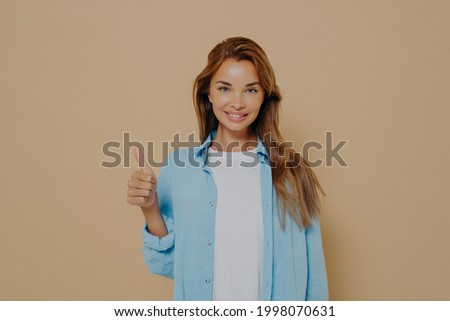 Happy young caucasian female in blue shirt with long sleeves rolled up weared over white tshirt making thumb up sign and smiling on camera with support on isolated pastel beige colored background