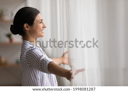 Overjoyed young Indian woman open curtains look in window distance meet welcome new day at home. Smiling millennial mixed race female feel excited about life career opportunities or perspectives. Royalty-Free Stock Photo #1998081287