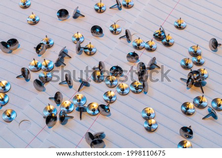 Background picture of numerous silver tacks scattered around white lined sheets of paper. The close up features several smooth top push pins lying around in several directions.
