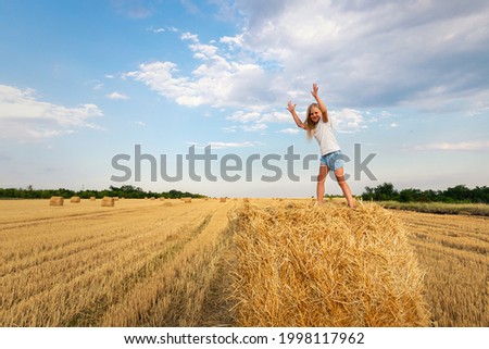 Portrait of cute little blond beautiful adorable cheerful caucasian kid girl enjoy sitting on hay stack or bale on harvested wheat field warm summer evening. Scenic rural country landscape background
