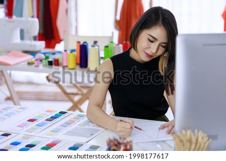 Smiling Asian woman dressmaker is sitting  sketch dress that she has to cut for the customer in the Tailor shop and sewing machines. Concept creating new clothes collection