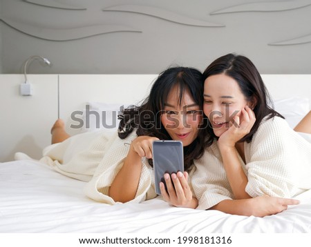 Two Asian women in bathrobes take a selfie with a mobile phone while lying in bed.
