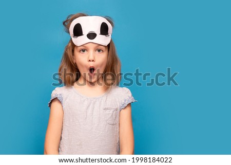 Portrait of young girl with surprised face and sleeping mask on head isolated over blue background. Empty space for text