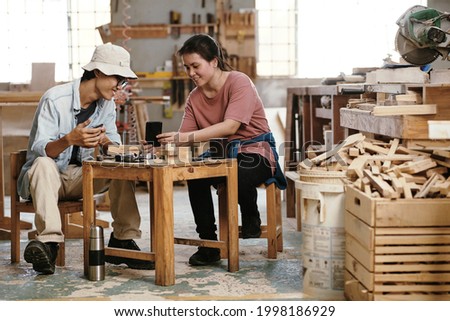 Joyful young carpenters enjoying coffee break in workshop, they are discussing picture of creative woodworking project on smartphones