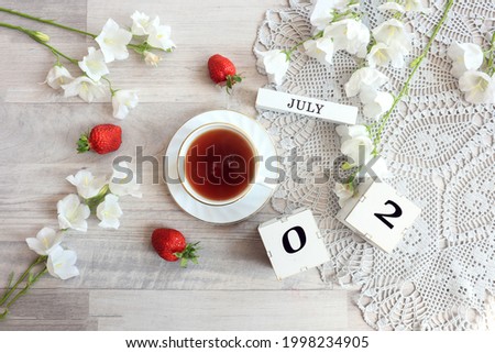 Calendar for July 2 : cubes with the numbers 0 and 2, the name of the month of July in English, a cup of tea on a gray openwork napkin, scattered flowers of bluebells and strawberries, top view