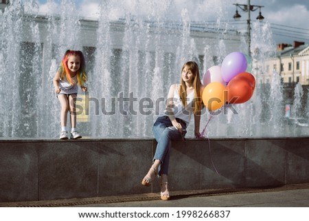 cute caucasian smiling girl with colorful dyed hair and her young mom holding baloons sitting on fountain and looking at her daughter going to jump. Image with selective focus 