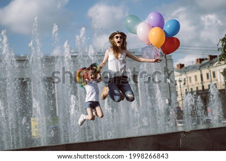 cute caucasian smiling girl with colorful dyed hair and her young mom holding baloons jumping from fountain. Image with selective focus 