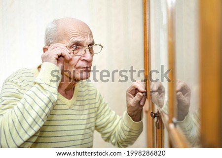 Senior male person holds eyeglasses with hand and open wooden door of bookcase in living room Royalty-Free Stock Photo #1998286820