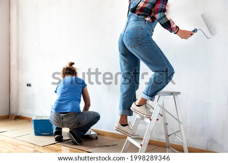 Two women painting apartment walls. Loading roller in paint tray. Using ladder to paint reach high. Renovating home. Real estate concept. Copy space. Royalty-Free Stock Photo #1998289346
