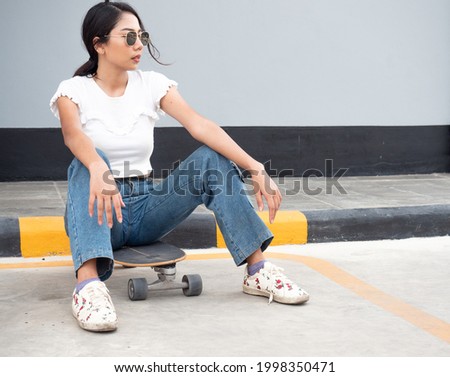 Exhausted woman skateboarder wears black eyeglasses sitting on board near pavement relaxing after surf skate exercising. Female hipster enjoy extreme sport. Trendy outdoor recreation in Thailand.