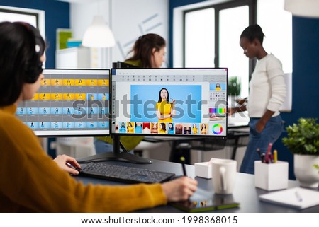 Woman retoucher with headset retouching photos using stylus pen drawing on graphic tablet, working on PC with two displays. Photo editor working in software app sitting in creative agency office
