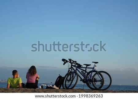 couple sits at a lake shore beach with bikes next to them, shot from the back. Space for text. Summer activities, relaxation, resting, relationship concept.