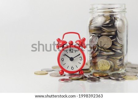 Selective focus of a red alarm clock and coins in a glass jar isolated on white background. Time management, financial, investment and saving money concept. This photo contains noise and grain.