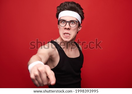 Funny young skinny guy in sportswear and nerdy glasses making angry face and outstretching hand towards camera against red background