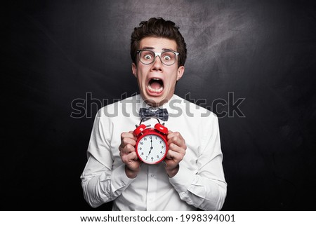 Young nerdy guy in formal outfit and eyeglasses showing alarm clock with 7 o clock time and screaming terrified while being late against black background