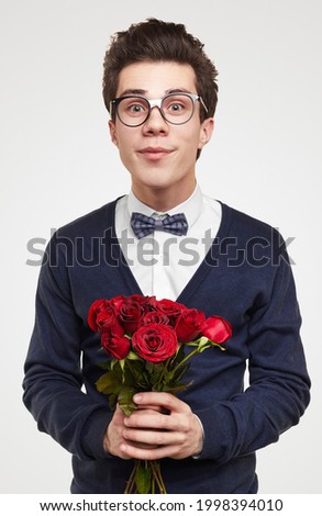 Young nerdy male in elegant suit and glasses with bouquet of red roses looking at camera against white background