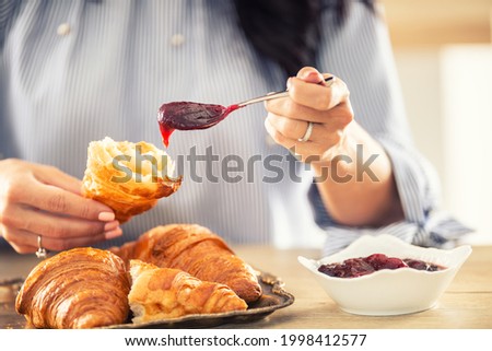 Female hands put jam onto a halved croissant in the morning. Royalty-Free Stock Photo #1998412577