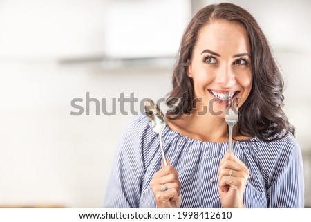 Woman thinking of eating something good bites into a fork while holding spoon in the other hand. Royalty-Free Stock Photo #1998412610