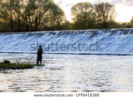 lonely silhouette of a fishing person on the river in the countryside with waterfall on background