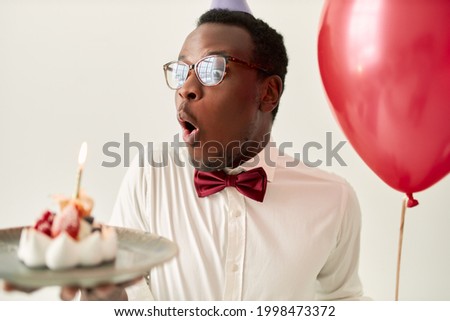Isolated image of funny fascinated young bearded dark skinned male wearing glasses and bow opening mouth, taking deep breath to blow out burning candle on birthday cake, making wish, holding balloon