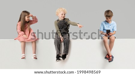 Three little kids, boys and girl sitting on box isolated over gray studio background. Copyspace for ad. Childhood, education, emotions, facial expression concept.