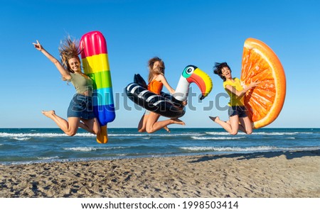 three happy girls on jumping motion with inflatable mattresses at the beach outdoors in summer celebrating vacation days. friends having fun on seaside holidays joy, carefree, genz and party concept Royalty-Free Stock Photo #1998503414