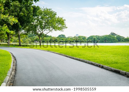 Empty street, green city park with blue sky. Pathway and beautiful trees track for running or walking and cycling relax in park on green grass field on the side. Sunlight and flare background concept Royalty-Free Stock Photo #1998505559