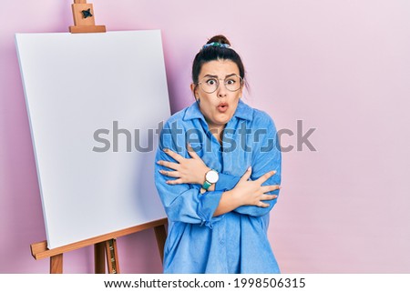 Young hispanic woman standing by painter easel stand shaking and freezing for winter cold with sad and shock expression on face 