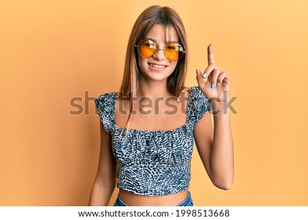 Teenager caucasian girl wearing fashion yellow sunglasses pointing finger up with successful idea. exited and happy. number one. 
