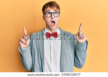 Young caucasian nerd man wearing glasses wearing hipster elegant look with bowtie amazed and surprised looking up and pointing with fingers and raised arms. 