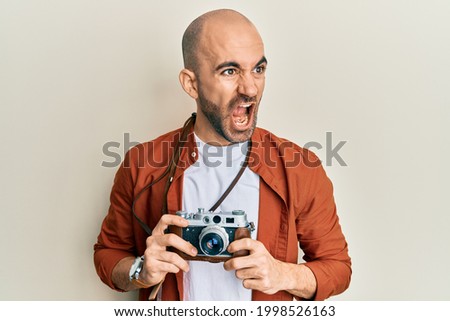 Young hispanic man holding vintage camera angry and mad screaming frustrated and furious, shouting with anger. rage and aggressive concept. 