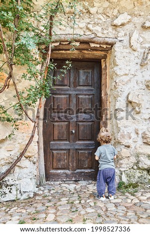 Back view of a lonely toddler standing outside in front of the closed wooden door of an old house. Concept of loneliness. Vertical photograph.