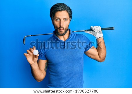 Young hispanic man holding golf ball making fish face with mouth and squinting eyes, crazy and comical. 