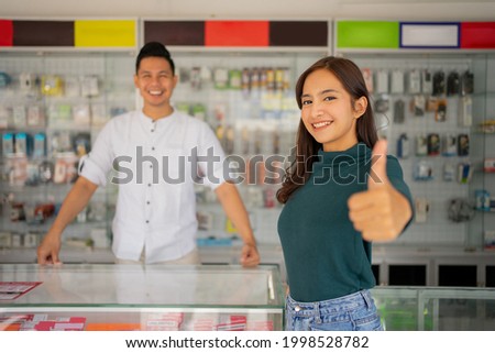 beautiful young woman with thumbs up standing against the background of a male shop assistant in a smartphone shop