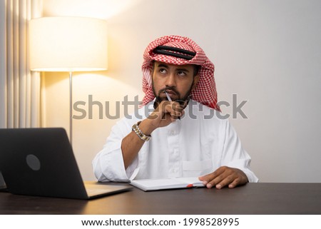Arab businessman in turban thinking while writing at work desk with office space background