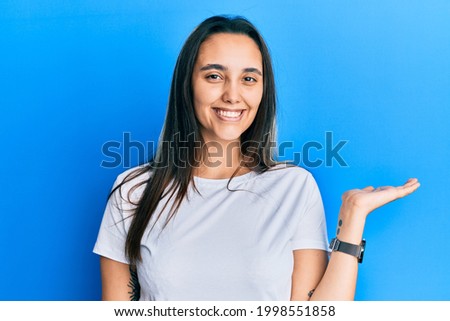 Young hispanic woman wearing casual white t shirt smiling cheerful presenting and pointing with palm of hand looking at the camera. 