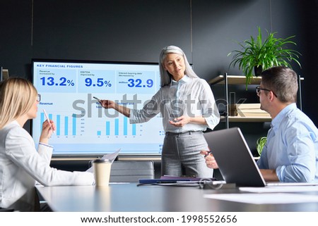Mid age senior Asian business woman ceo executive manager showing to colleagues team income revenue data figures planning on big screen in modern office on business seminar workshop.
