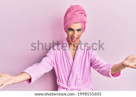 Young man wearing woman make up wearing shower towel on head and bathrobe smiling cheerful offering hands giving assistance and acceptance. 