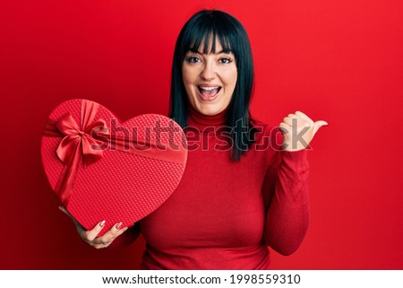 Young hispanic woman holding valentine gift pointing thumb up to the side smiling happy with open mouth 