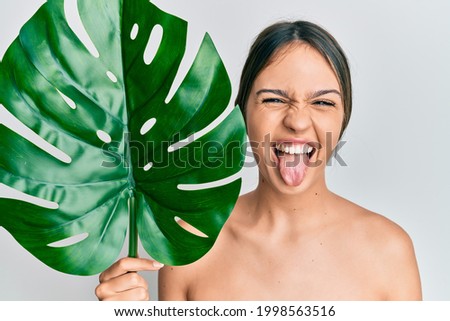 Young brunette woman holding green plant leaf close to beautiful face sticking tongue out happy with funny expression. 