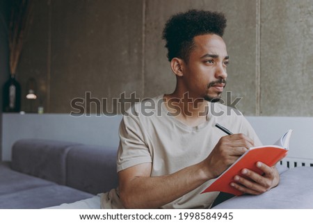 Pensive thoughtful young poet student african american man 20s wearing beige t-shirt sitting on grey sofa indoors apartment write down memories in notebook diary, resting on weekends staying at home. Royalty-Free Stock Photo #1998584945