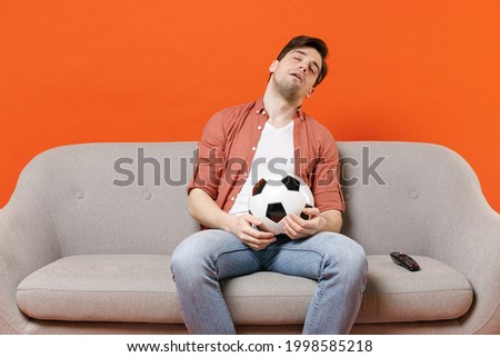 Young tired man football fan in shirt support team sit on sofa home watch tv live stream hold soccer ball sleeping isolated on orange background studio portrait People lifestyle sport leisure concept Royalty-Free Stock Photo #1998585218