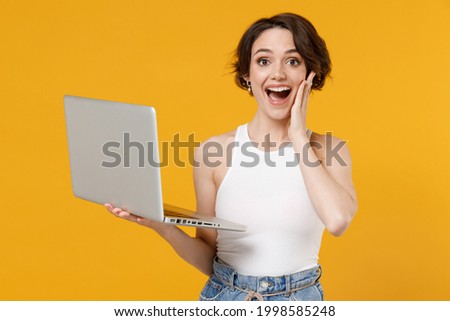 Young freelancer surprised happy woman 20s with bob haircut wearing white tank top shirt using laptop pc computer chat online browsing surfing internet hold face isolated on yellow color background